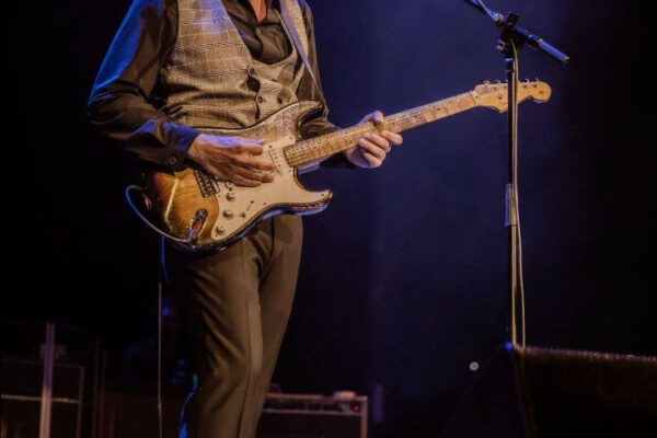 Male guitarist on stage singing into a microphone while playing a sunburst electric guitar on a dimly lit stage.