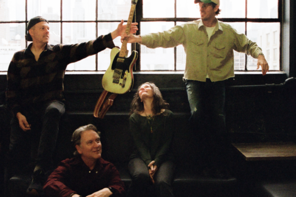 Four people in a loft pass a white electric guitar between two men while a woman sits on steps and looks upward.