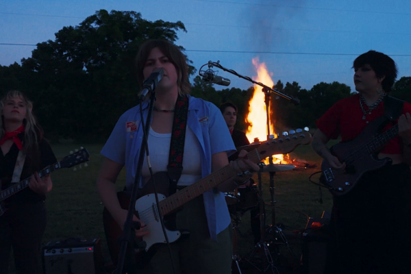A four-member rock band performing outdoors at dusk around a large bonfire, with a female lead singer playing an electric guitar and singing into a microphone.