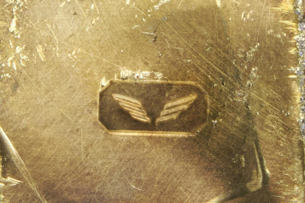 Brass panel with a small hexagonal badge showing a pair of stylized wings, surface worn and scratched.
