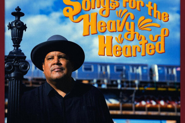 Chris Pierce album cover: man in dark clothing and hat beside a decorative lamppost, bold orange title 'Songs For The Heavy Hearted' against a blue sky and train backdrop.