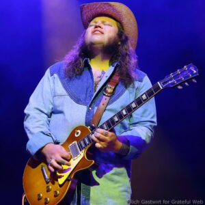 Guitarist wearing a denim shirt and brown hat, playing a sunburst electric guitar on stage with blue lighting.] ,