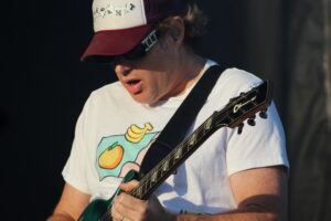 Guitarist wearing a white shirt with a fruit graphic, a red and white cap, and sunglasses, playing a green electric guitar on stage.