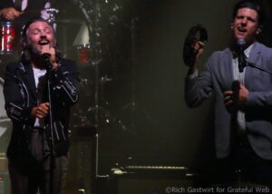 Two male vocalists perform on stage: left singer with eyes closed, wearing a black embroidered jacket and singing into a microphone; right singer in a gray suit with a tambourine and microphone.