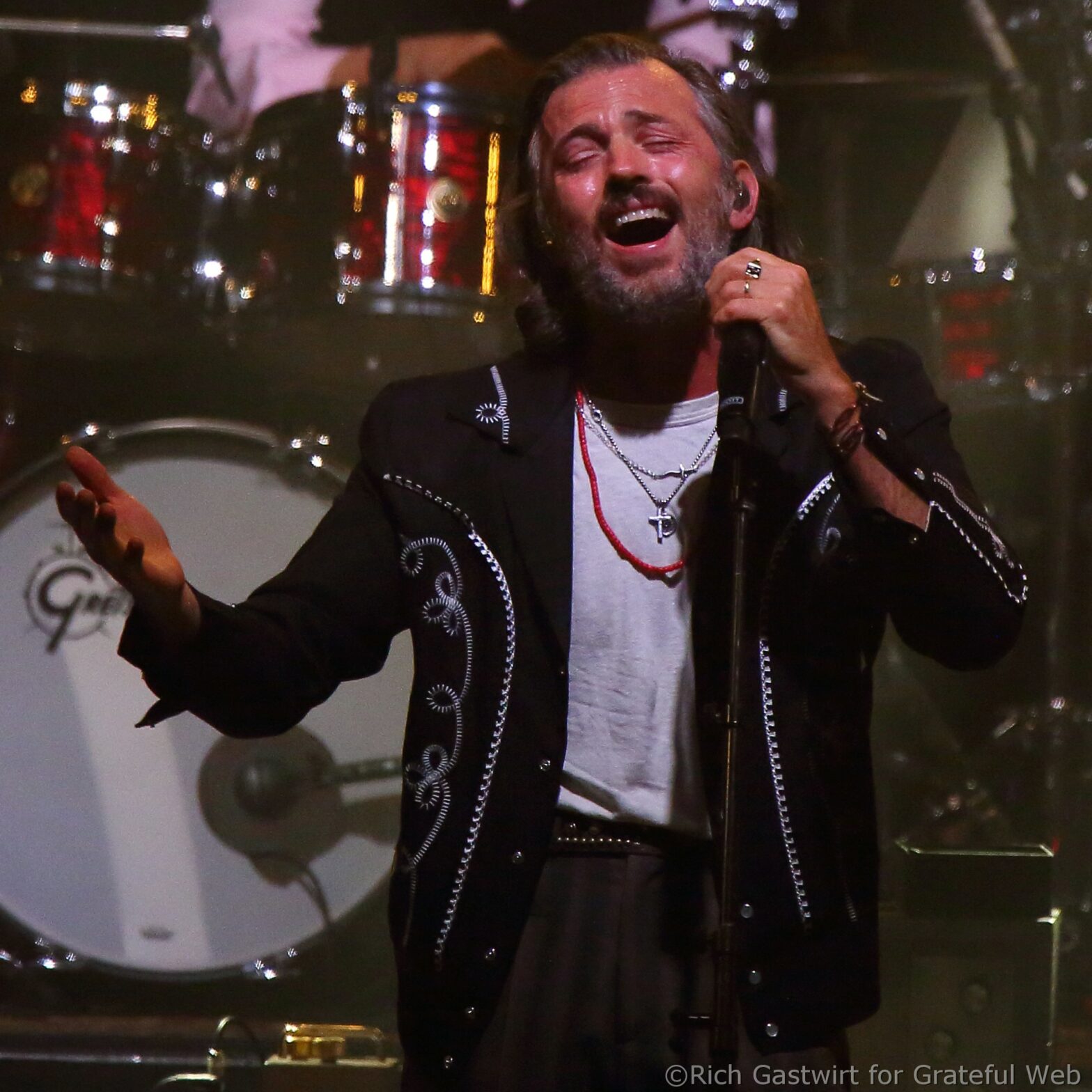 Male singer with gray beard, eyes closed, passionately singing into a microphone on stage, wearing a black embroidered jacket and white shirt with layered necklaces.