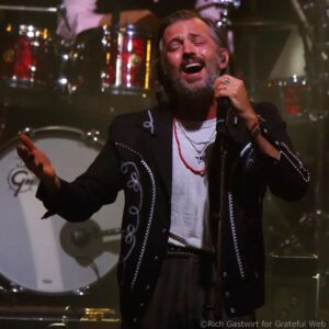 Male singer with gray beard, eyes closed, passionately singing into a microphone on stage, wearing a black embroidered jacket and white shirt with layered necklaces.