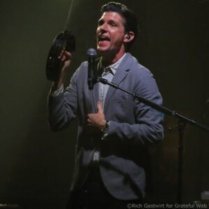 Male singer in a light gray blazer performing on stage, singing into a microphone while holding a tambourine in his left hand.