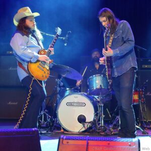 Two guitarists on stage performing, one wearing a cowboy hat and denim, the other in a denim shirt playing electric guitars with a drummer visible in the background.