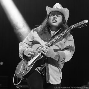 Musician wearing a cowboy hat and denim shirt plays an electric guitar on stage with eyes closed.