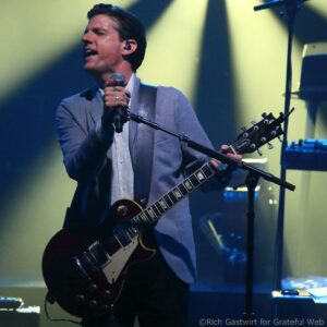 Male guitarist singing into a microphone on stage with a sunlit yellow and blue light backdrop, holding a dark Les Paul-style guitar.