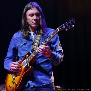 Female guitarist in a denim shirt playing a sunburst Gibson Les Paul on stage, focused on the fretboard.
