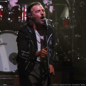 Male vocalist with closed eyes singing into a microphone on stage, wearing a black embroidered jacket.