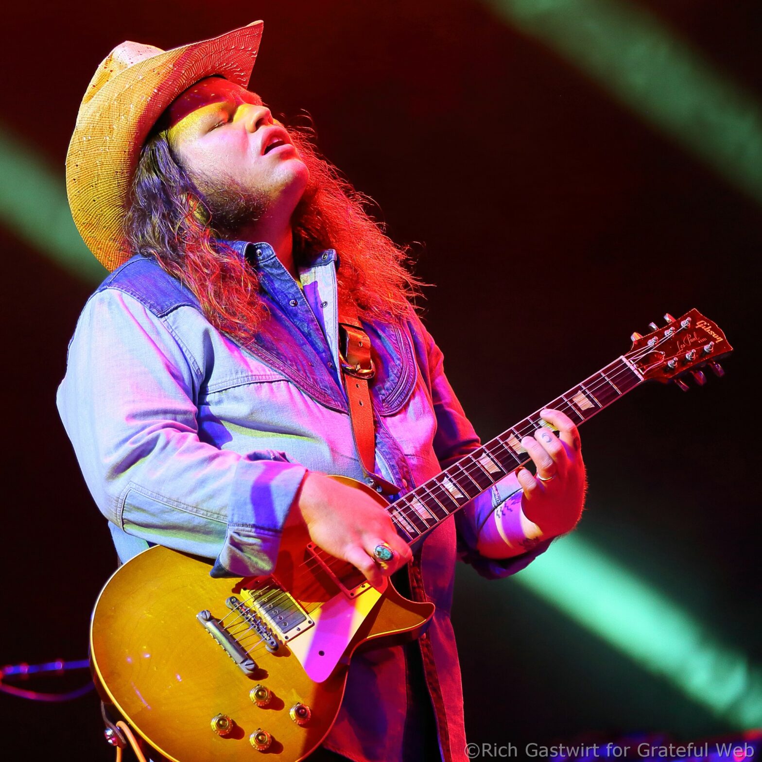 Musician wearing a straw hat and blue denim shirt, playing a yellow Gibson-style electric guitar on stage with colorful stage lighting