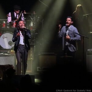 Two male singers perform on stage with microphones, drummer visible at back behind a drum kit amid hazy stage lighting.
