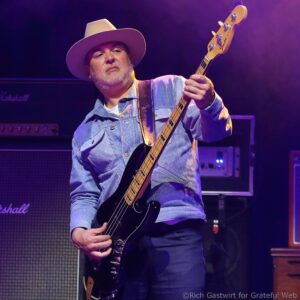 Musician wearing a wide-brimmed hat plays a black bass guitar on stage with purple lighting.
