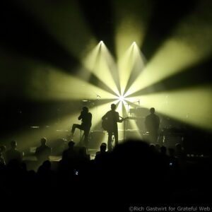 Concert stage with bright yellow spotlights forming a star-like pattern behind silhouette of musicians and instruments.