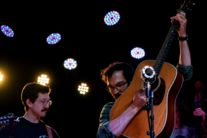 Guitarist raises his acoustic guitar toward the microphone on a dark stage with colorful circular lights in the background, another musician visible on the left.