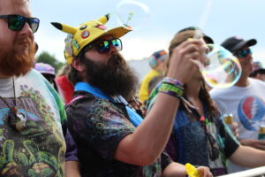 People at an outdoor festival blowing bubbles; man with red beard and sunglasses in foreground wearing a tie-dye shirt, others in colorful hats nearby.