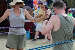 Two people hula-hooping outdoors at a sunny festival, with onlookers in the background.