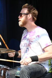 Bearded man wearing sunglasses plays drums at an outdoor event.