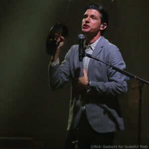 Male singer on stage, wearing a gray blazer, singing into a handheld microphone on a dimly lit set.
