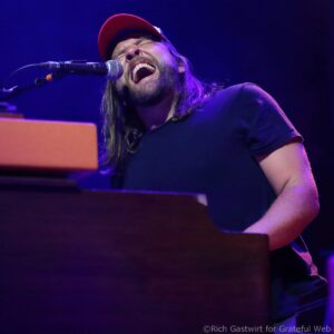 Male singer wearing a red cap, eyes closed, passionately singing into a microphone on a dimly lit stage.