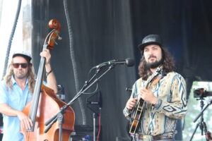 Two musicians on stage: a guitarist singing into a microphone and a double bassist wearing a cap and sunglasses while playing.