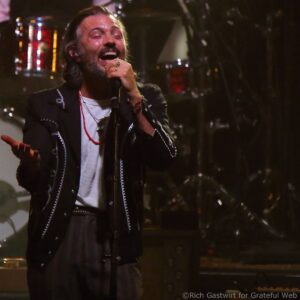Male singer with a beard passionately singing into a microphone on a dimly lit stage, eyes closed.
