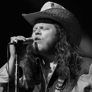 Close-up of a singer wearing a woven cowboy hat, eyes closed, singing into a microphone on stage.