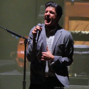 Male singer in a gray blazer singing into a handheld microphone on a dimly lit stage, eyes closed in performance.