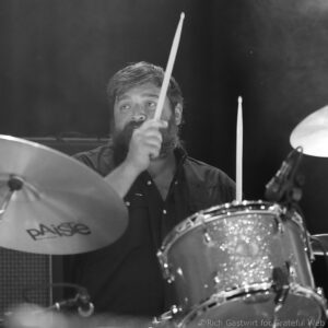 Close-up of a drummer mid-stroke, stick raised above a cymbal in a black-and-white concert photo.