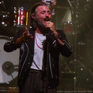 Male singer with eyes closed, passionately singing into a microphone on stage, wearing a black embroidered jacket and silver necklaces.