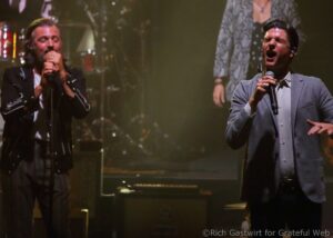 Two male singers perform on stage, each holding a microphone, one in a black jacket on the left and the other in a light gray blazer on the right, with stage lights and a keyboard visible in the background.