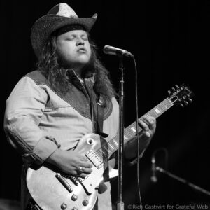 Male guitarist singing into a microphone while wearing a cowboy hat on stage, playing an electric guitar (black and white).