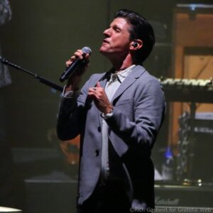 Male singer in a light blazer performs on stage, holding a microphone close to his mouth with his eyes closed.