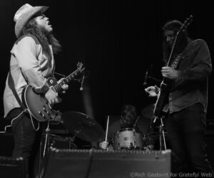 Live concert scene with two guitarists facing each other: left musician in a cowboy hat and denim, playing a Les Paul-style guitar; right musician with long hair in a denim jacket playing another guitar. A drum kit is visible in the background.