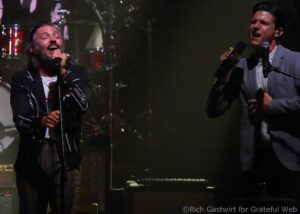 Two male singers performing on stage with microphones; left singer sings with eyes closed, right singer holds a tambourine and sings into a mic.