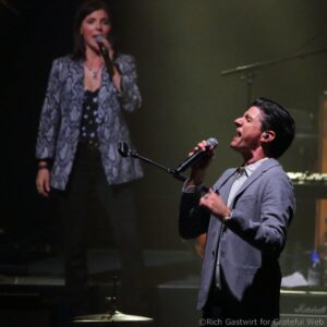 Stage performance with a female vocalist on the left wearing a patterned jacket, singing into a microphone, and a male vocalist in a suit on the right with a microphone as well.