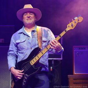 Male bassist wearing a light denim shirt and a wide-brim hat performs on stage, hands on a black Fender Jazz Bass under purple stage lighting.
