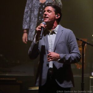Male singer on stage, wearing a light gray blazer and white shirt, singing into a handheld microphone with an in-ear monitor visible in his ear.