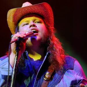 Singer wearing a straw hat, close-up while singing into a microphone on a colorful stage.