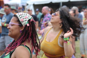 Two women dancing at an outdoor festival, wearing colorful outfits and accessories, with a blurred crowd in the background