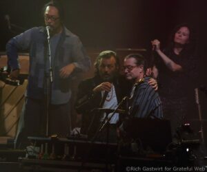 Stage photo of a live band: a man with a microphone at center-left, another guitarist seated in front, and a female singer on the right with a microphone.