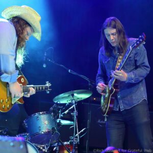 Two guitarists performing on a blue-lit stage: one in a straw hat on the left and a denim-clad player on the right with a guitar.