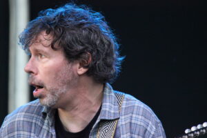 Close-up side profile of a man with gray stubble wearing a blue plaid shirt, on a stage with a guitar neck visible on the right edge.