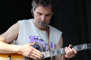 Man playing an acoustic guitar, wearing a sleeveless shirt, with a blue guitar pick in his mouth and a focused expression.