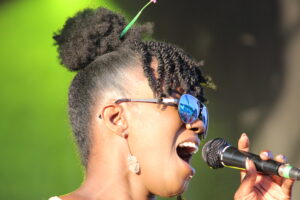 Close-up of a female singer with braided hair and blue mirrored sunglasses singing into a microphone against a green background.