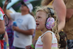 Girl with rainbow-pattern headphones in a side profile at an outdoor event, glitter on her cheeks.