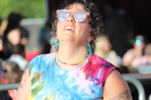 Woman outdoors wearing pink sunglasses, turquoise earrings, and a colorful tie-dye shirt at a sunny festival.