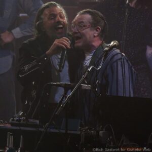 Two male singers perform on a dimly lit stage, singing into microphones with a keyboard visible in the foreground.